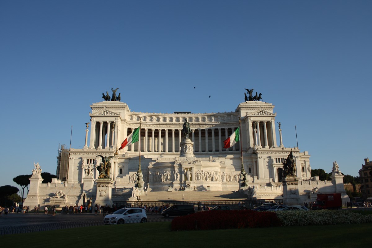 An ornate marble building with bronze statues of angels and chariots, and the Italian flag on either side.